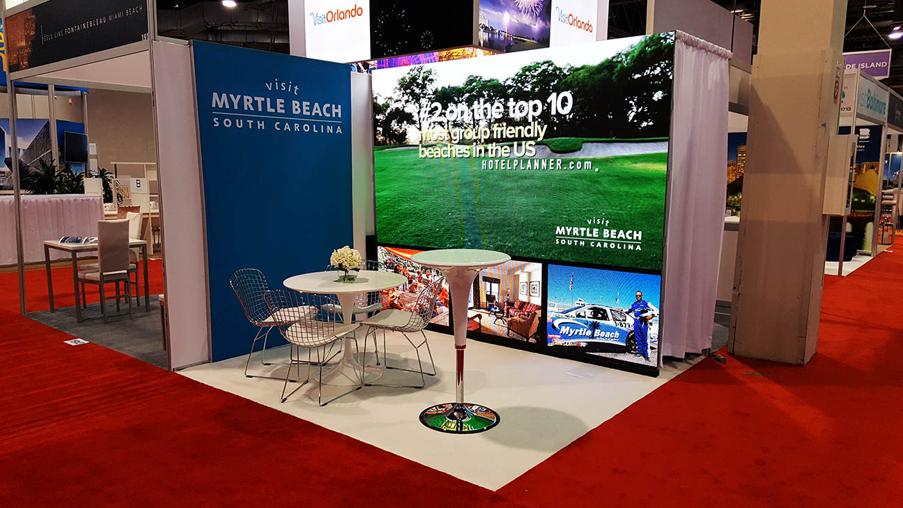 10x10 trade show booth with 10x8 LED video wall, fabric side panel, table, four chairs, and visitor greeting area